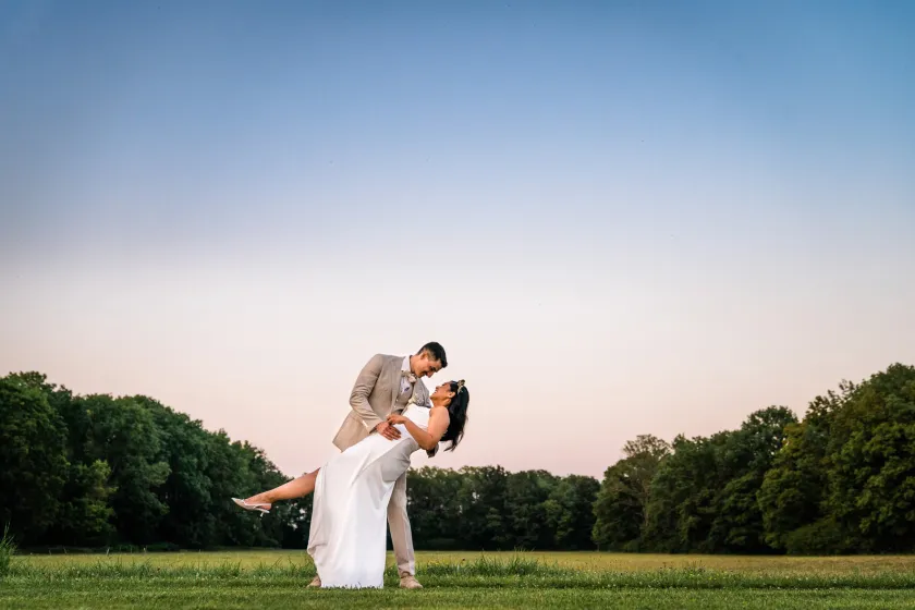 Ein Foto, dass den Bräutigam zeigt, wie er die Braut in seinen Armen hält und sie nach hinten kippt, während die Sonne am Horizont untergeht.
